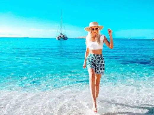 A guest wading through clear shallow water on a white pebble beach, a catamaran anchored offshore