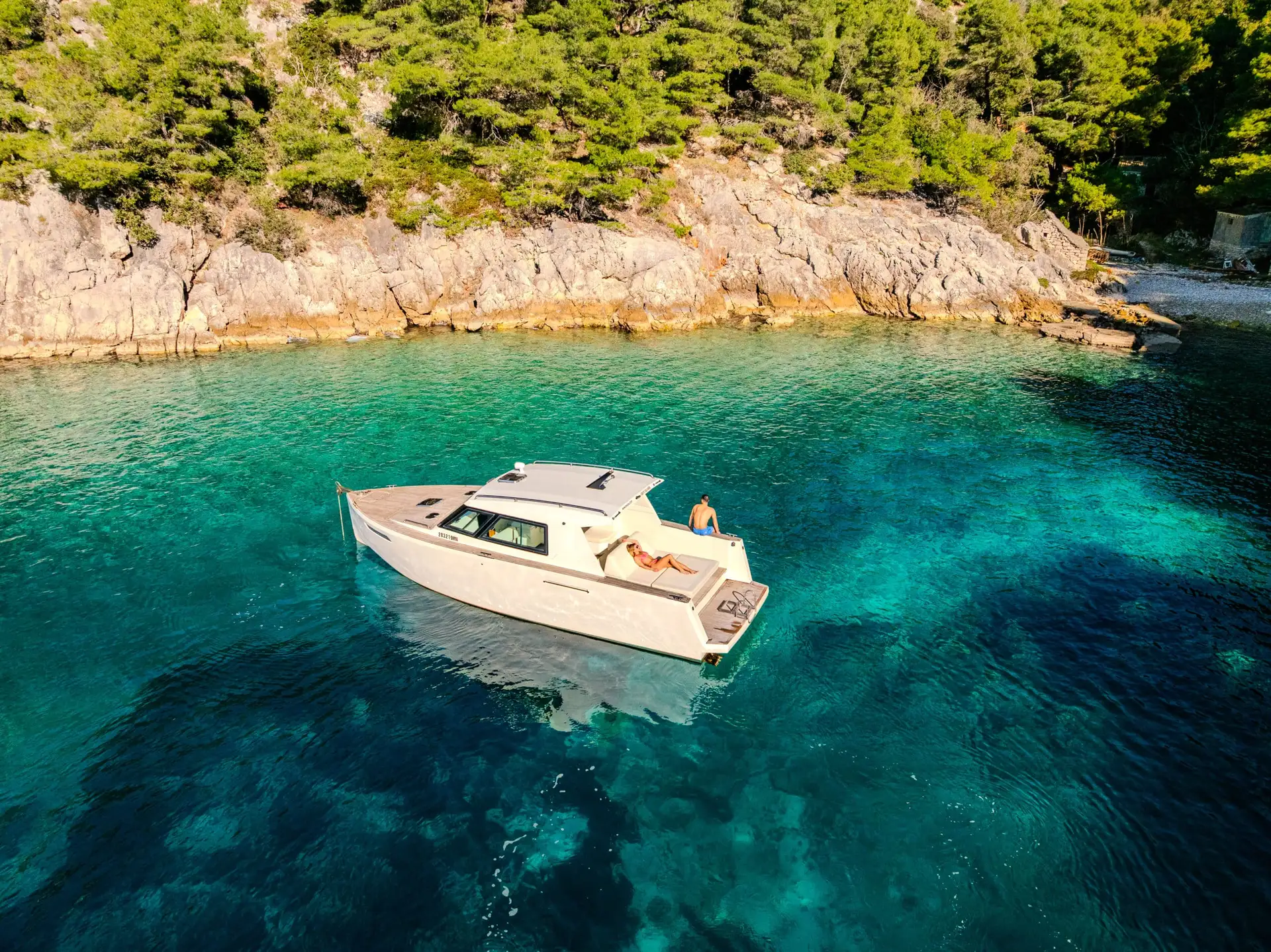 A private boat anchored in a turquoise bay below a pine-covered rocky shore on the Adriatic coast